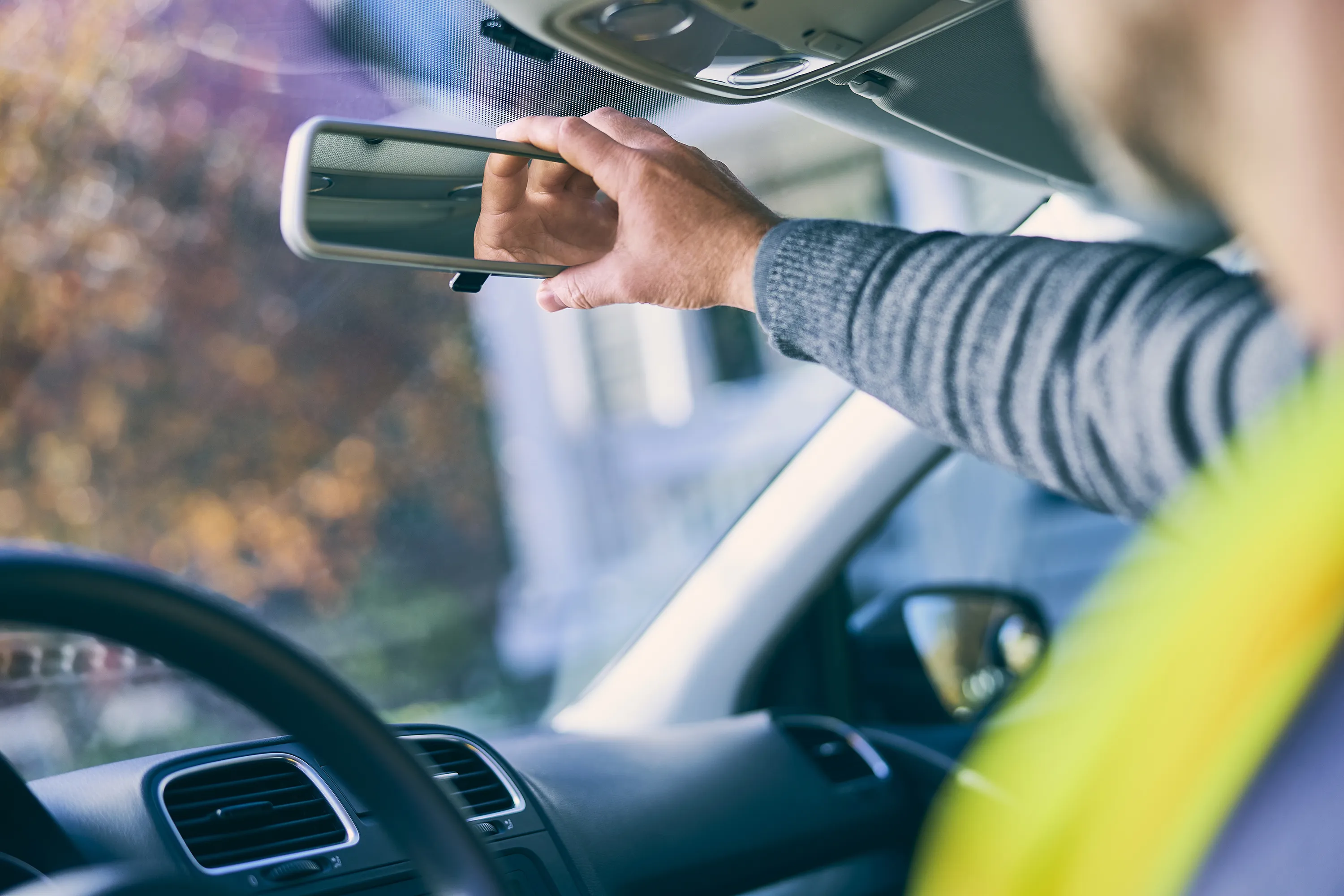 An individual in a green vest sits in the driver seat of a vehicle and adjusts the rearview mirror.