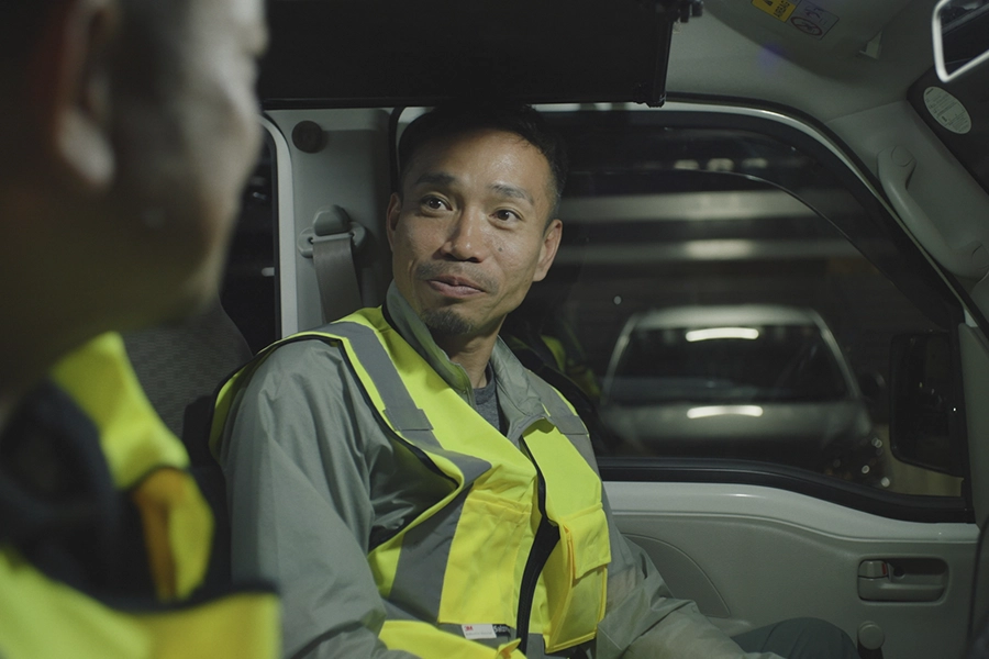 Man in a yellow vest sitting in a car looking at another man in a yellow vest.